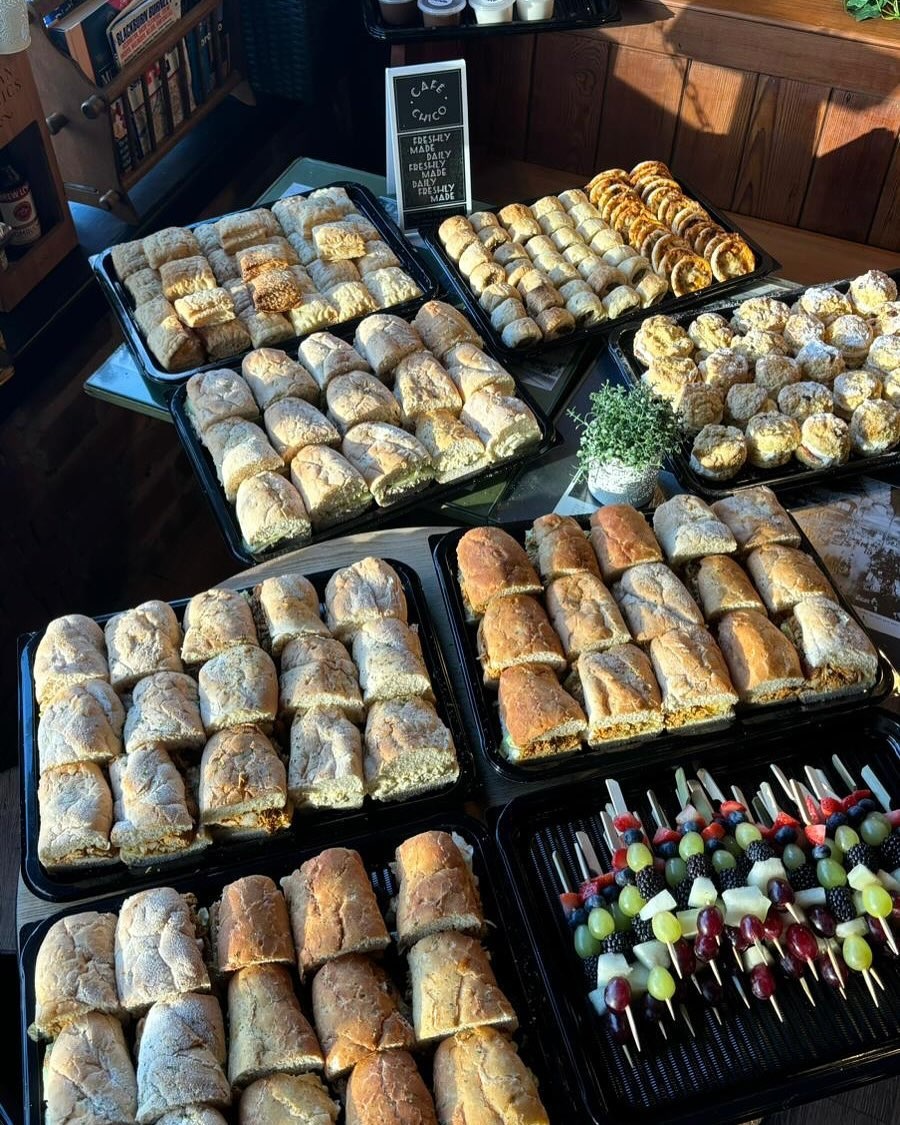 Café Chico real bread & sandwich platter — rustic bread loaves, ciabatta sandwich rolls, cookies and fruit skewers on black trays, with a Café Chico chalkboard sign reading 'Freshly Made Daily'